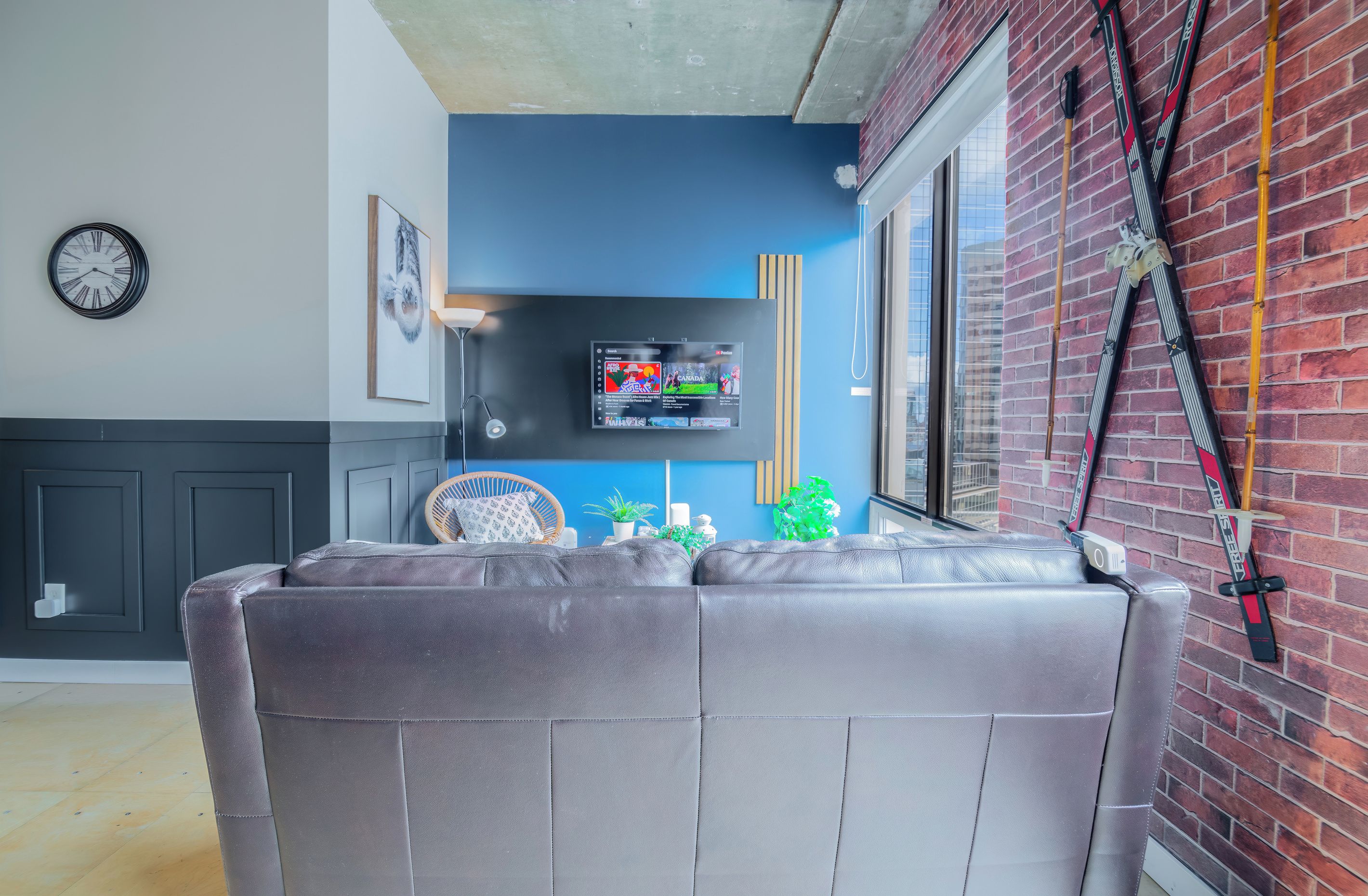 Un salon moderne avec un canapé gris, un téléviseur fixé au mur, une horloge et un porte-skis adossé à un mur en briques.
