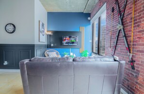 A modern living room with a grey sofa, a mounted TV, a clock, and a ski rack against a brick wall.