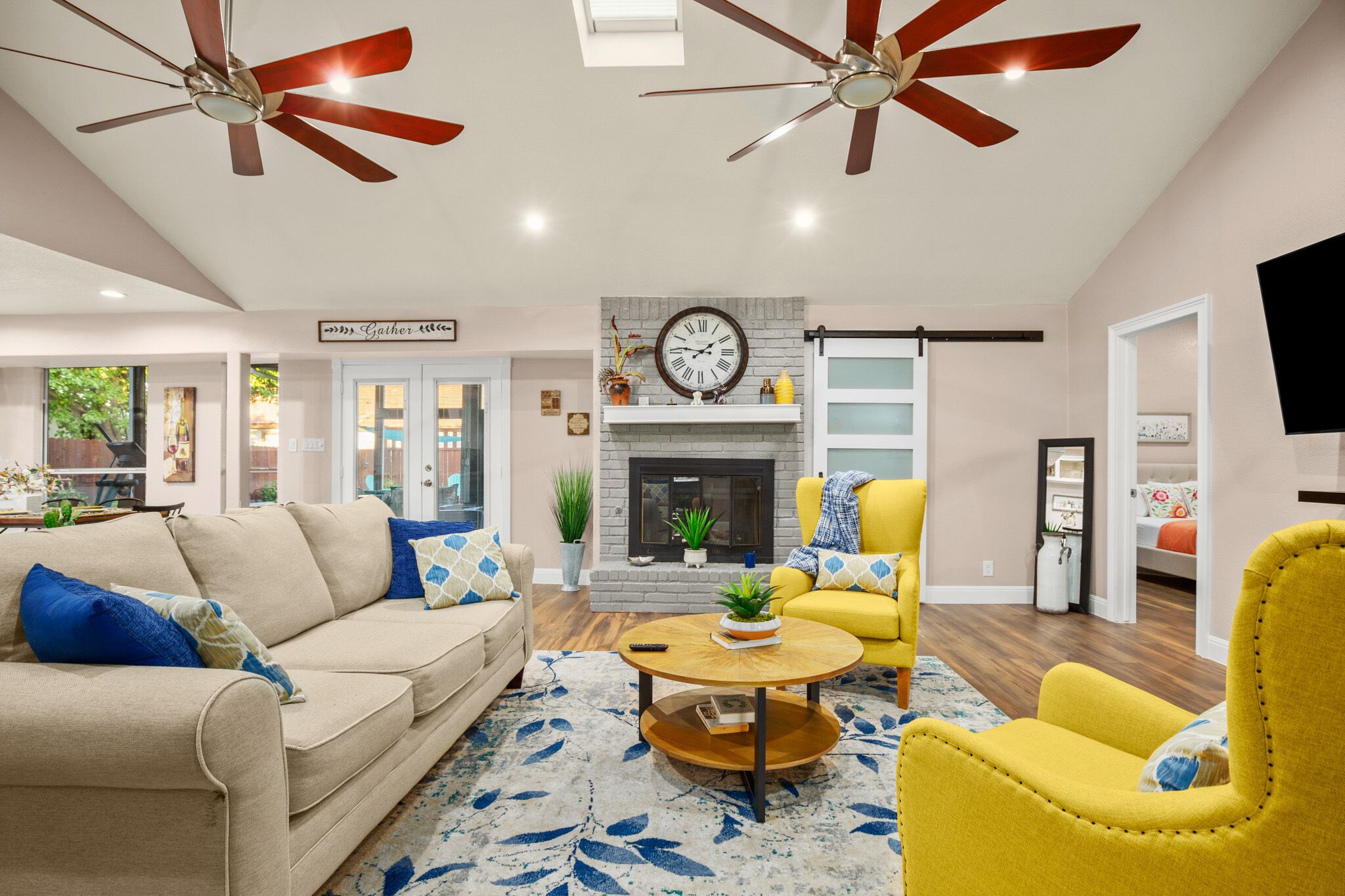 A spacious living room with a beige sofa, yellow armchair, and a fireplace. There are ceiling fans, a clock, and a TV mounted on the wall.