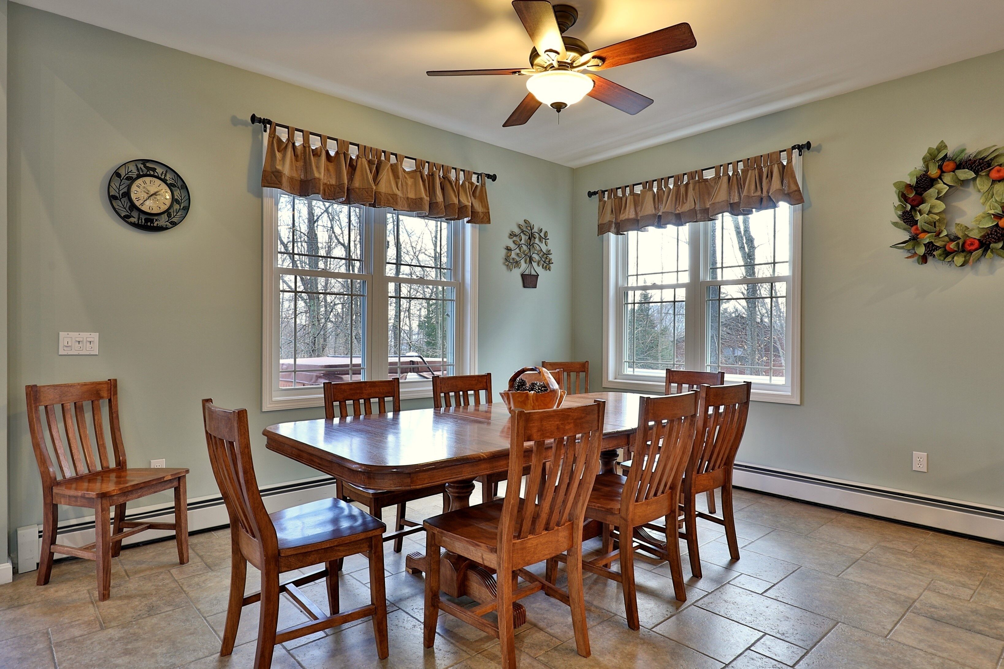 A dining room with a large wooden table, six chairs, a ceiling fan, and two windows with curtains.