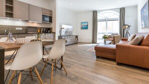 A modern living room with a brown leather sofa, a dining area with a wooden table and white chairs, and a kitchen with beige cabinets and a large window. - Exklusiv. Strandnah. Zentral (Westerland)