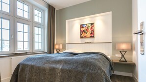 A bedroom with a bed, two bedside tables with lamps, a window with curtains, and a painting on the wall. - Exklusiv. Strandnah. Zentral (Westerland)