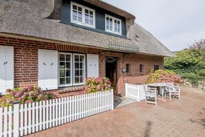 Une maison traditionnelle en briques, avec un toit de chaume, des volets blancs et une clôture en bois peinte en blanc.