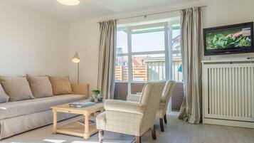 Ein Wohnzimmer mit einer Couch, Sesseln, einem Couchtisch, einem Fernseher und einem Fenster mit Vorhängen.