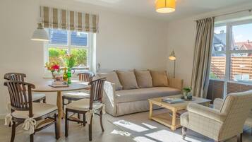 Ein gemütliches Wohnzimmer mit einer Couch, einem Couchtisch und Sesseln. Es gibt einen Esstisch mit Stühlen und ein Fenster mit Vorhängen.