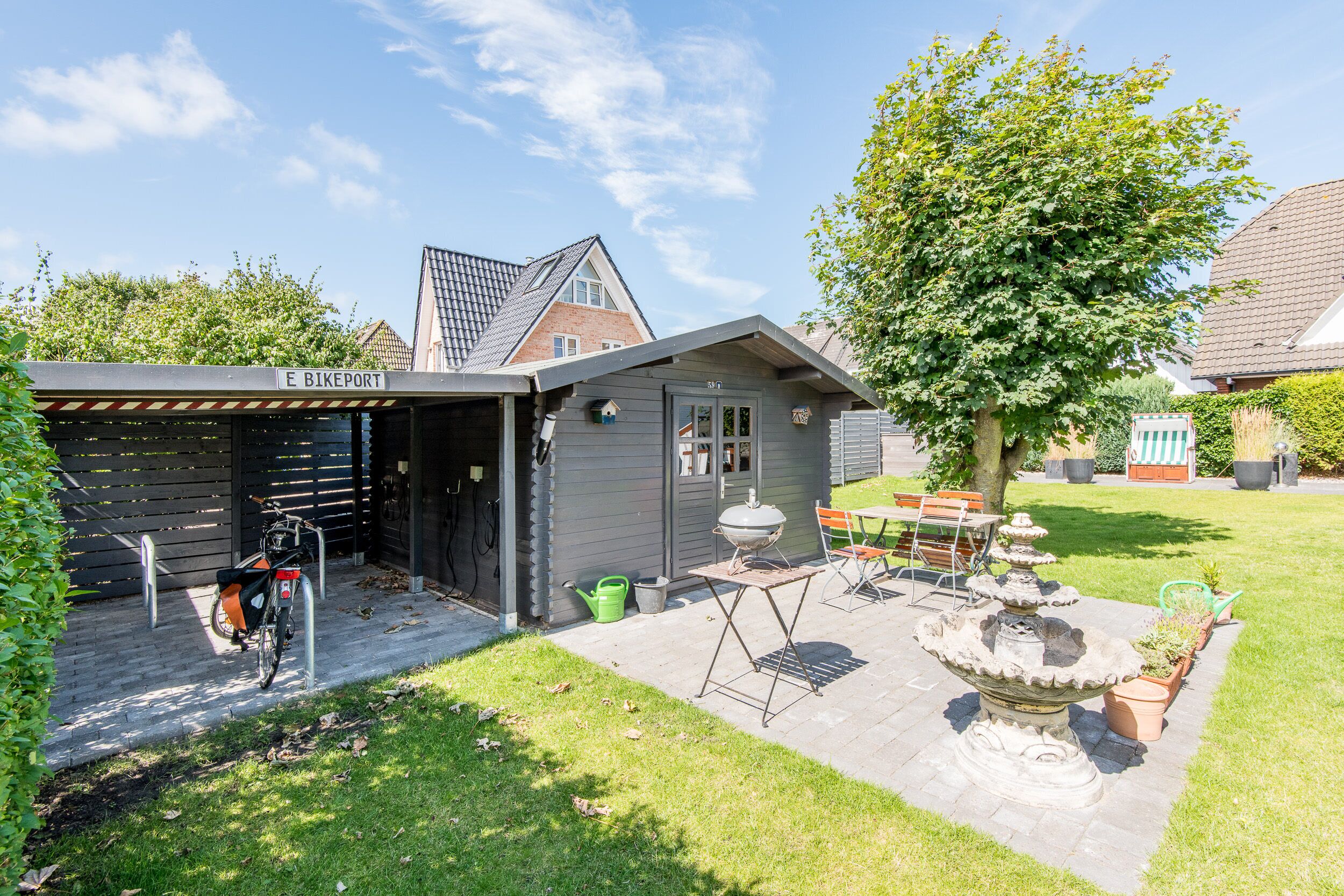 Une maison avec un garage fermé portant l’inscription « E BIKEPORT », un barbecue, un vélo et une fontaine en pierre dans la cour.
