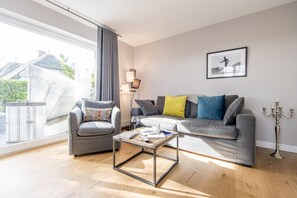 A modern living room with a grey sofa, a matching armchair, a coffee table, and a large window offering a view of a building outside.