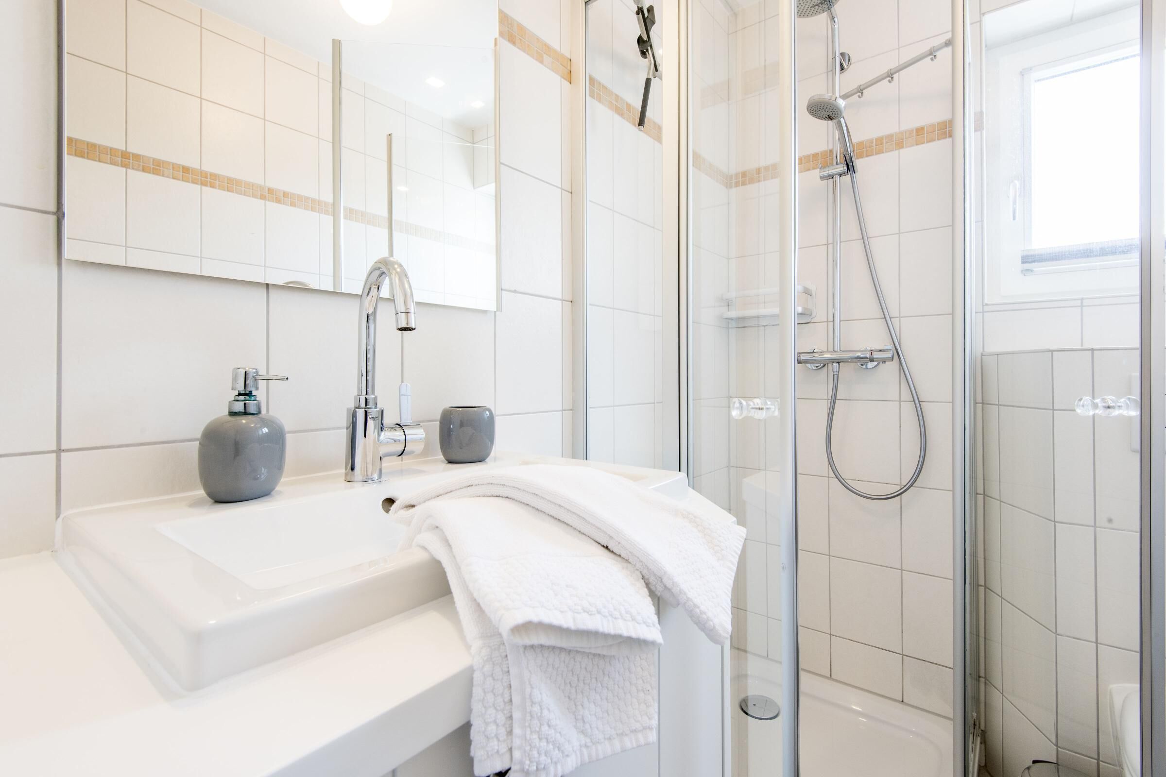Ein modernes Badezimmer mit einem weißen Waschbecken, einem Spiegel, einer Dusche mit Glastür und einem Fenster für natürliches Licht.