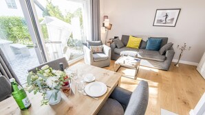 A modern living room with a dining table set for two, a grey sofa with cushions, and a large window offering a view of the outdoors. - Modern. Ruhig. Südterrasse (Westerland)