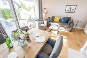A modern living room with a dining table set for two, a grey sofa with cushions, and a large window offering a view of the outdoors.