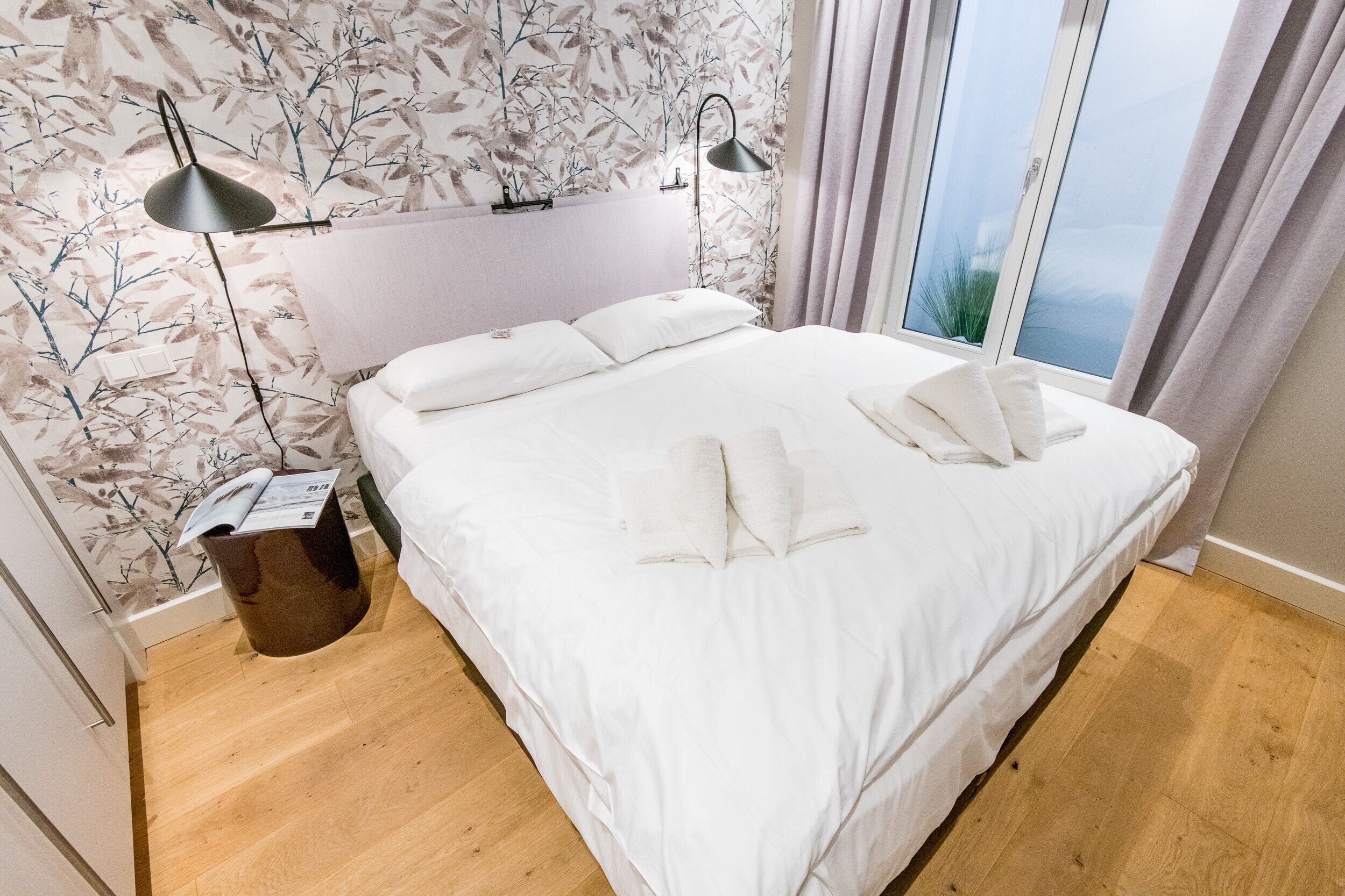 A neatly made bed with white linens and pillows, a bedside lamp, and a wooden floor.