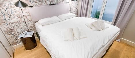 A neatly made bed with white linens and pillows, a bedside lamp, and a wooden floor.