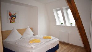 Ett sovrum med en säng, vita sängkläder, ett träpanelat tak, ett fönster med växter och en färgstark tavla på väggen.