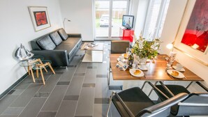 A modern living room with a sofa, dining table, and chairs. There is a television on a stand and a window with a view of the outside. - Hell. Entspannend. Komfortabel (Westerland)