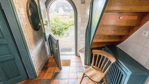 A narrow hallway with a wooden chair, a door leading outside, and a view of a house through an archway.