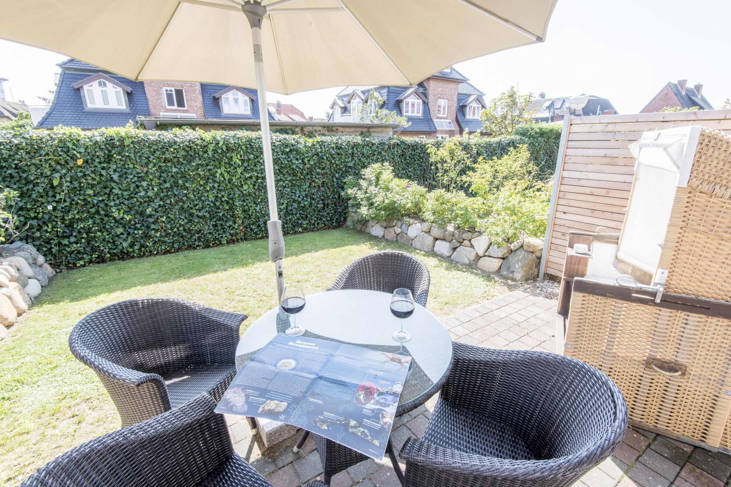An outdoor patio with wicker furniture and a table, surrounded by a well-maintained hedge and a stone wall.