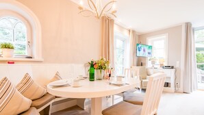 Een eetgedeelte met een ronde tafel, beiges bekleden stoelen, een kroonluchter en een televisie op de achtergrond.