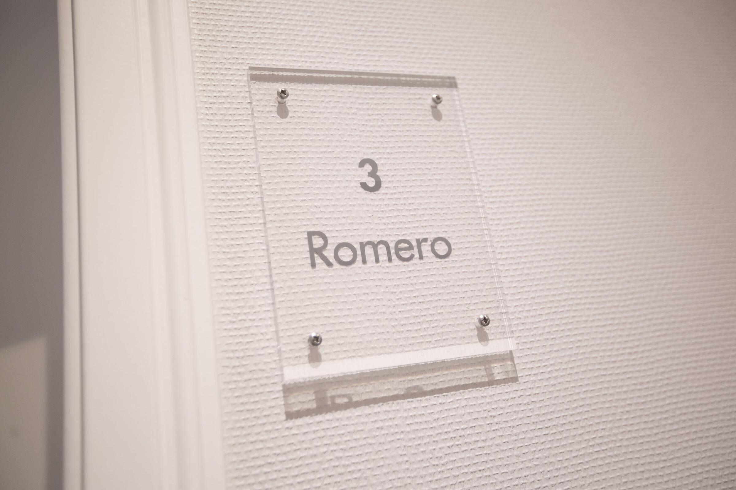 Ein Schild mit der Zimmernummer 3 und dem Namen Romero an einer strukturierten Wand.