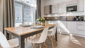 A modern kitchen with white cabinets, a wooden dining table, and a large window with blinds. - Art your Life - Residence, Whg. 3 Romero (Westerland)