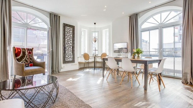 A modern living room with a round glass coffee table, a sofa with patterned cushions, a dining area with a table and chairs, and large windows with curtains. - Art your Life - Residence, Whg. 3 Romero (Westerland)