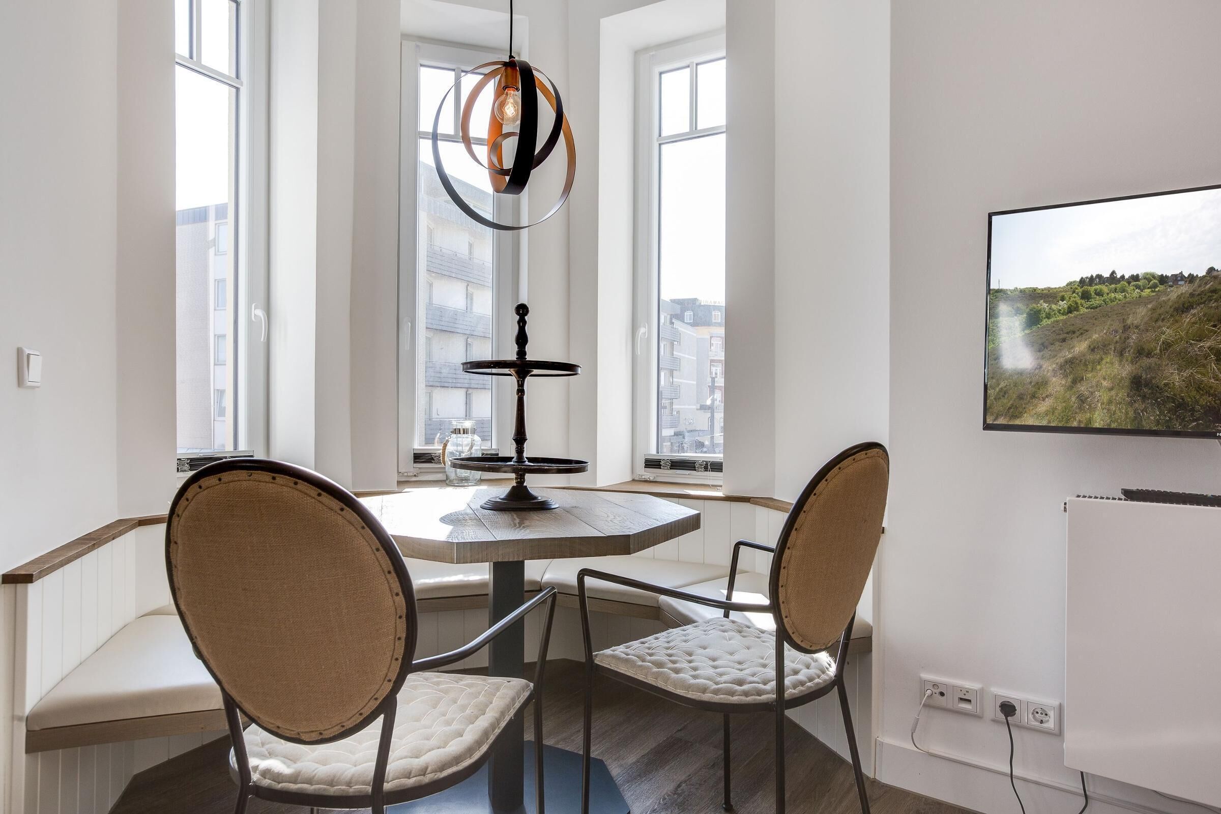 A modern dining area with a round table, two chairs, a pendant light, and a TV on the wall.