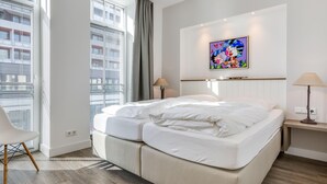 A modern hotel room with a large bed, a desk, and a chair. There is a window with blinds and a painting on the wall. - Art your Life - Residence, Whg. 3 Romero (Westerland)