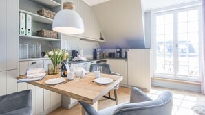A modern kitchen with white cabinets, a wooden dining table, and a large window providing natural light. - Exquisit. Wellness. Balkon. Höög Hüs, Whg. 2A Objekt-id: Whw/02a (Westerland)