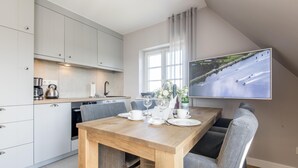 A modern kitchen with white cabinets, a wooden dining table set for two, and a large flat-screen TV mounted on the wall displaying a scenic river view. - Komfortabel. Ruhig. Gemütlich (Westerland)