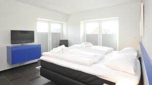 Une chambre d’hôtel moderne avec un lit, un téléviseur posé sur une commode bleue et de grandes fenêtres.