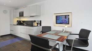 Une cuisine compacte avec des meubles blancs, un four à micro-ondes et une machine à café. On y trouve une table à manger avec deux chaises, ainsi qu’un tableau abstrait encadré accroché au mur.