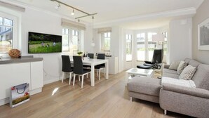 A modern living room with a grey sofa, a dining area with a white table and chairs, and a kitchen area with a TV mounted on the wall. - Strandnah. Ruhig. Komfortabel. Objekt-id: Hdr/04 (Westerland)