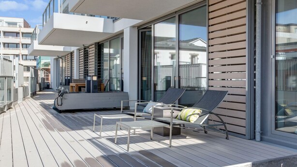A modern outdoor deck with lounge chairs and a table, large sliding glass doors, and a view of neighboring buildings. - Modern. Apart. Lichtdurchflutet. Neue Mitte, Whg. 4 Objekt-id: Nmw/04 (Westerland)