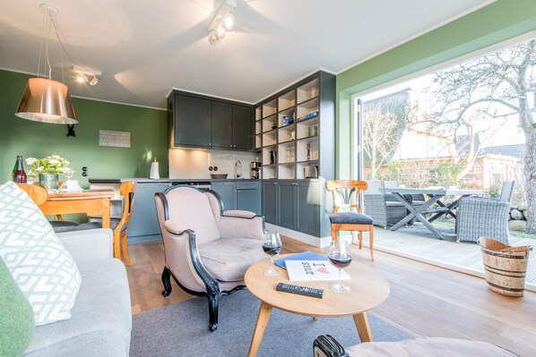 Ein modernes Wohnzimmer mit Sofa, Sessel und Couchtisch. Eine Küche mit Schränken sowie ein Essbereich mit Tisch und Stühlen sind zu sehen.