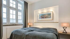 A neatly made bed with a textured blanket, a framed cityscape painting on the wall, and two bedside lamps on a wooden nightstand. - Strandnah. Meerblick. Wellness (Westerland)