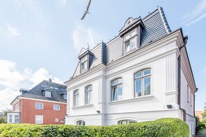 Un bâtiment à deux étages, doté d’une façade blanche et d’un toit gris, avec de nombreuses fenêtres et une cheminée bien visible.