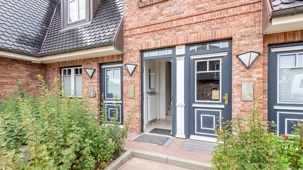 Een huis van baksteen met een donkerblauwe deur en raamkozijnen, een kleine tuin met groene planten en een pannendak.