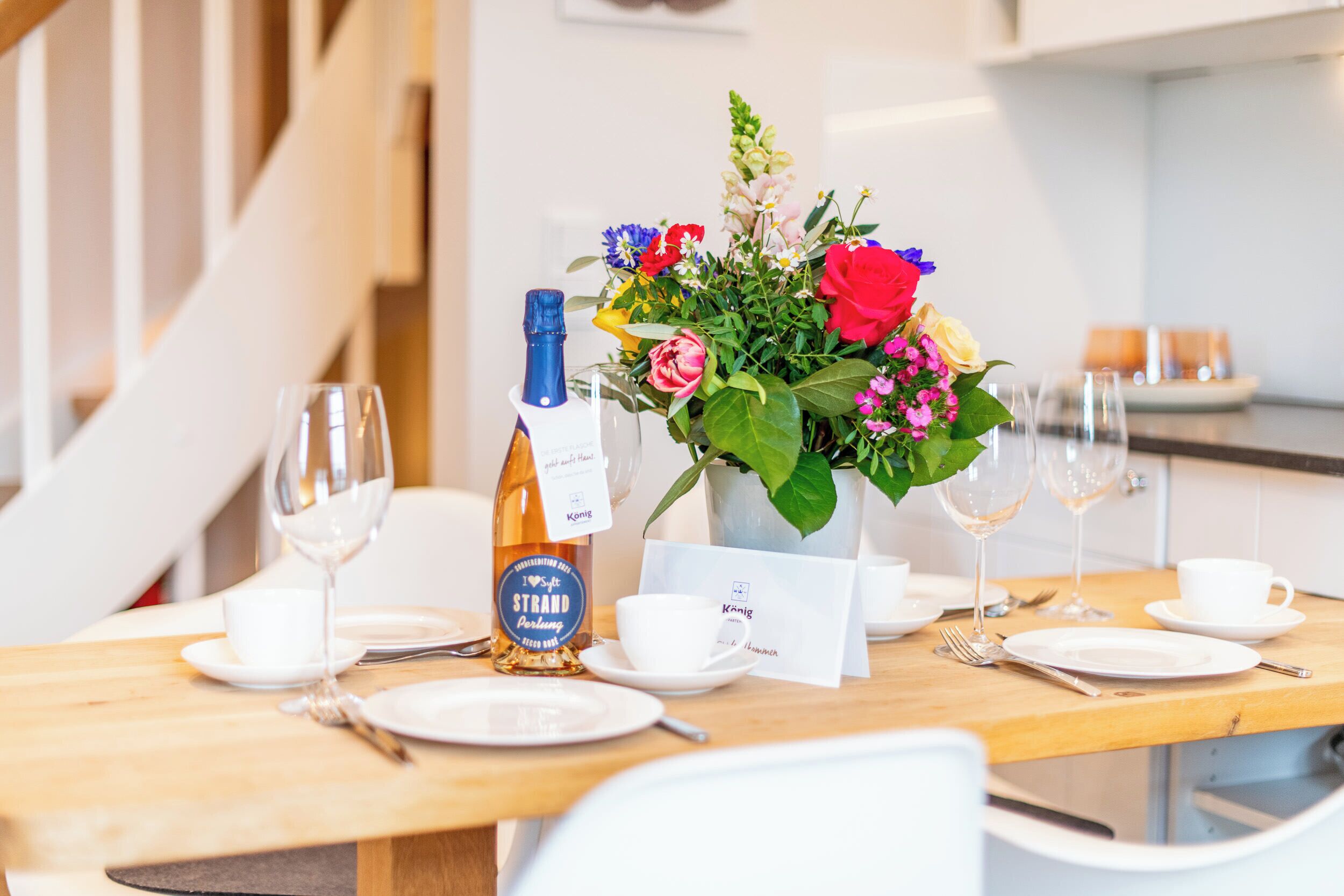 A dining table set with plates, cutlery, a bottle of wine, and a floral centerpiece.