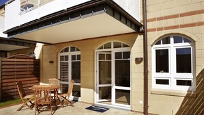 A two-story building with a covered patio area, featuring a wooden table and chairs, and a sliding glass door.