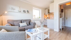 Ein modernes Wohnzimmer mit einem grauen Sofa, einem weißen Couchtisch und einer Zeitschrift. Im Hintergrund ist eine Küche mit weißen Schränken und einem Fenster erkennbar.
