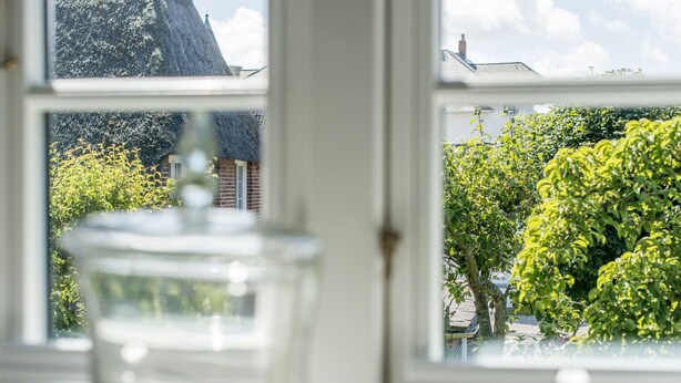 A view through a window with a clear glass vase in the foreground, a brick building, and a tree with green foliage. - Sonnenfreisitz. Geschmackvoll. Exklusiv (Westerland)