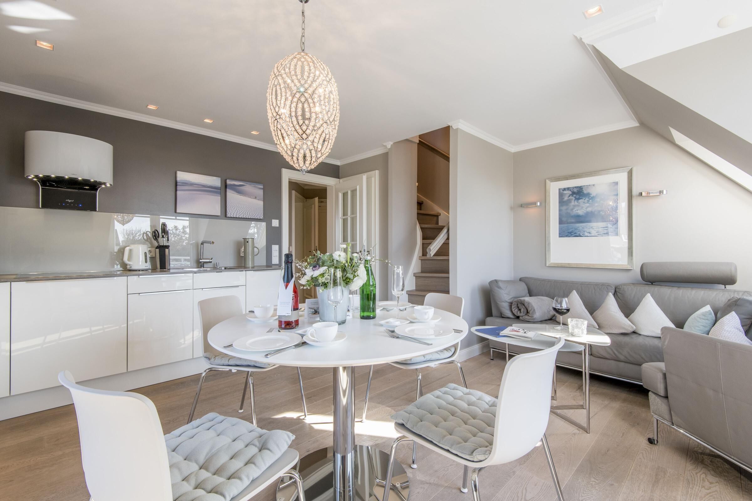 Eine moderne Küche mit weißen Schränken, ein Essbereich mit einem runden Tisch und ein graues Sofa in einem großzügigen Wohnzimmer.