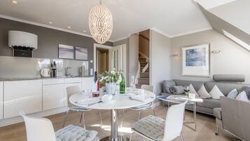 Eine moderne Küche mit weißen Schränken, ein Essbereich mit einem runden Tisch und ein graues Sofa in einem großzügigen Wohnzimmer.