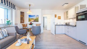 A modern kitchen with white cabinets, a dining area with a wooden table and chairs, and a living area with a sofa and a window. - Zentral. Strandnah. Gemütlich. Haus Hauke, Whg. 8 Objekt-id: Hh/08 (Westerland)