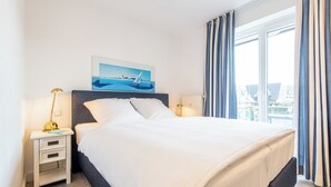 A hotel room with a bed, a nightstand with drawers, a lamp, a painting of a sailboat, and a window with curtains. - Zentral. Strandnah. Gemütlich. Haus Hauke, Whg. 8 Objekt-id: Hh/08 (Westerland)