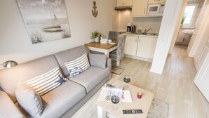 Ein modernes Wohnzimmer mit einem grauen Sofa, einem Couchtisch mit einer Zeitschrift, Weingläsern und einer Fernbedienung. Im Hintergrund ist eine Küche mit weißen Schränken und einer Mikrowelle zu sehen.