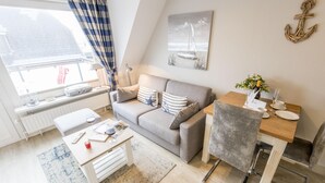 Ein gemütliches Wohnzimmer mit einer Couch, einem Esstisch und einem Sessel. Es gibt ein Fenster mit Vorhängen, eine Lampe und ein Bild an der Wand.