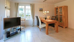 A modern living room with a flat-screen TV, a dining table with chairs, a cabinet with glass doors, and a window with curtains. - Exklusiv. Modern. Zentral. Haus Nordmarkgarten, Whg. 7a Objekt-id: Hng/07a (Westerland)
