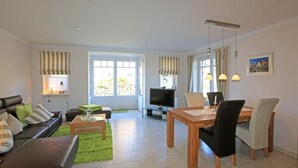 A living room with a sofa, coffee table, TV, and dining area with chairs and a table. - Exklusiv. Modern. Zentral. Haus Nordmarkgarten, Whg. 7a Objekt-id: Hng/07a (Westerland)