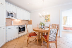 A modern kitchen with white cabinets, a built-in microwave, and an oven. There is a dining table with chairs and a chandelier above.