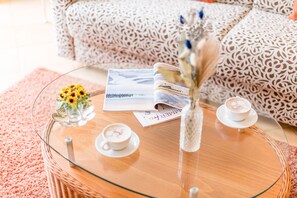 A glass-top coffee table with a wicker base, a vase with dried flowers, two saucers with cups of coffee, and a magazine. - Schwimmbad. Ostbalkon. Zentral. Residenz Bismarck, Whg. 8 Objekt-id: Hrb/08 (Westerland)